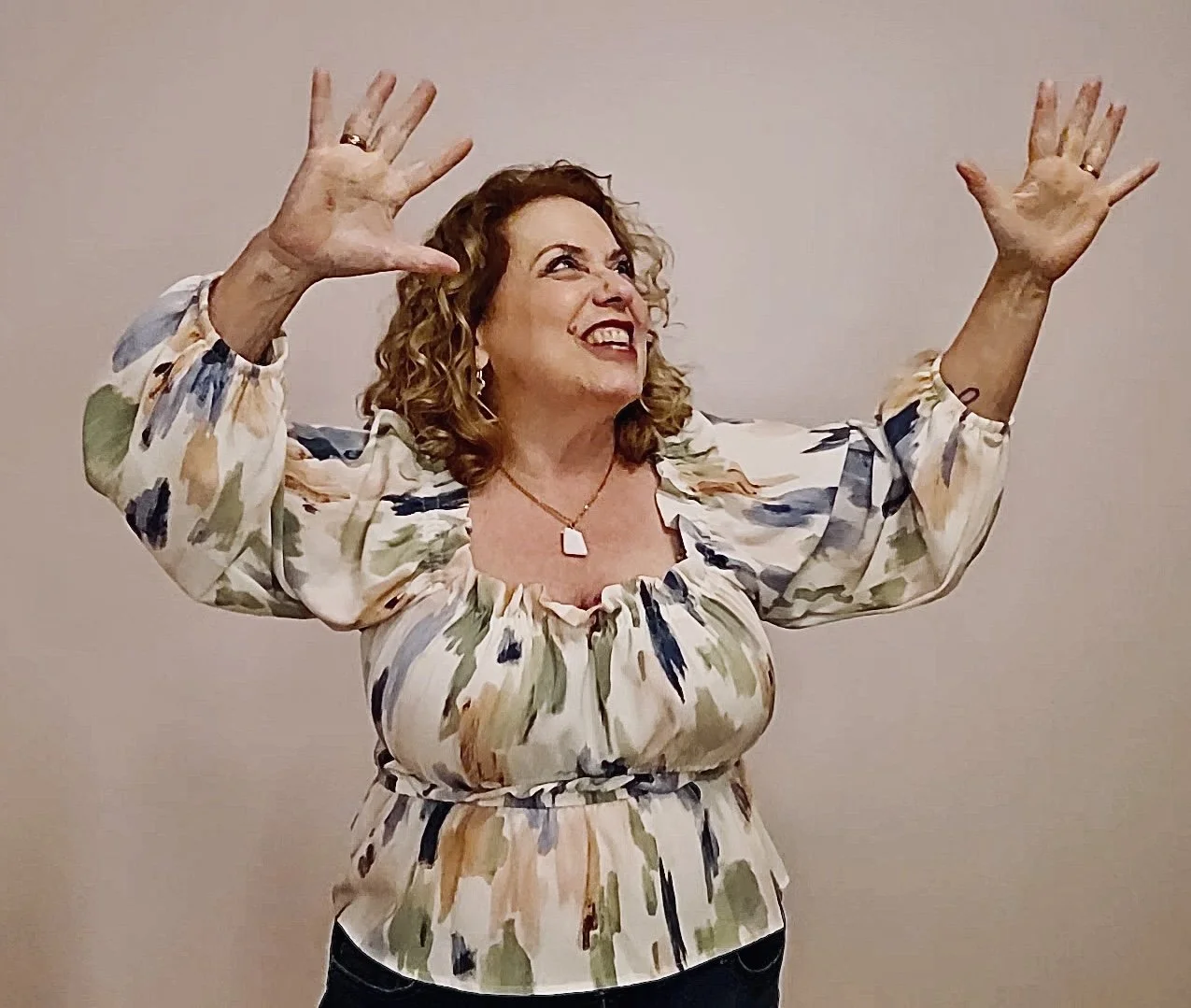 A woman with curly hair smiling and raising her hands up, wearing a floral blouse and jewelry, standing against a plain background.