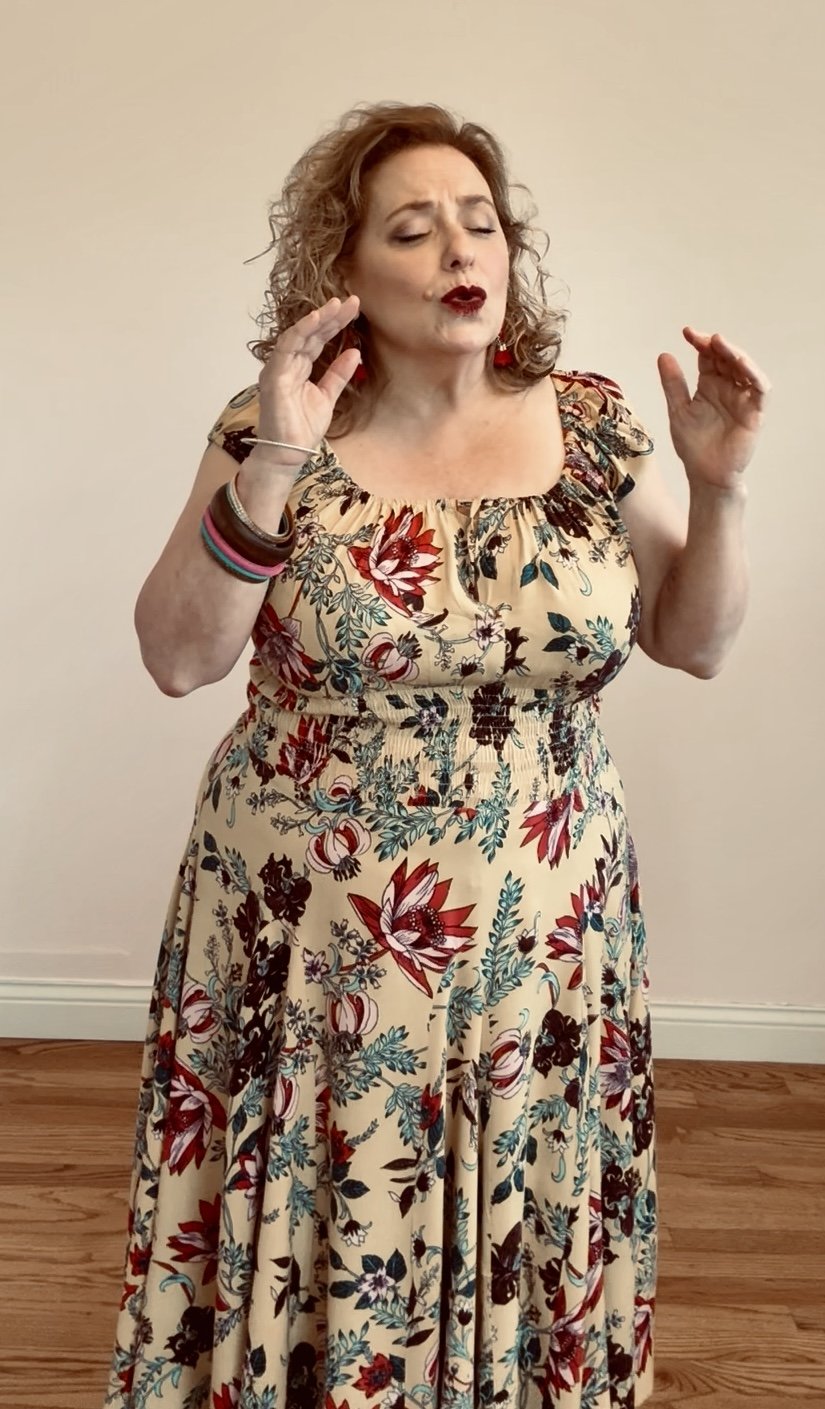 Kitty Karn, in a scene where a woman with curly hair wearing a floral dress, with her eyes closed and hands raised near her face, expressing emotion.
