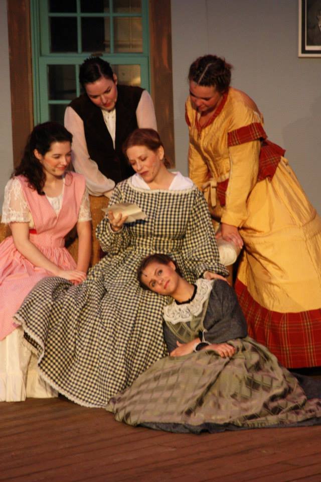 Kitty Karn in Five women dressed in period costumes are gathered around one woman who is lying down, reading a book, in a cozy indoor setting.