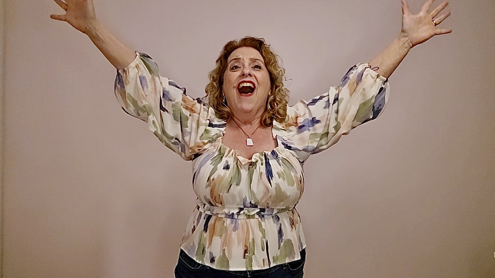 Woman with curly hair wearing a floral blouse with arms raised and mouth open as if excited or shouting