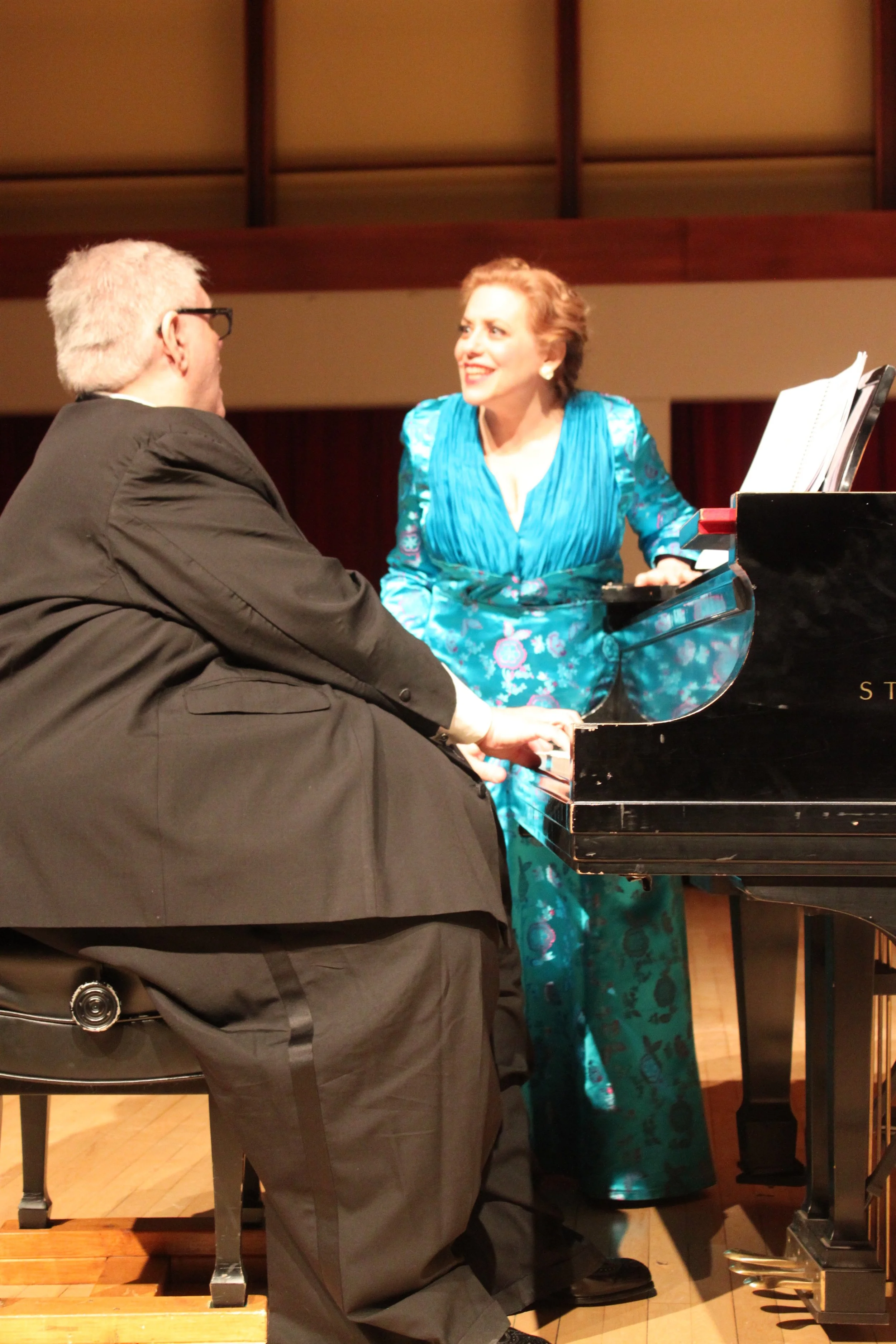 Two people sitting and playing a black grand piano, one man with gray hair and glasses, and a woman in a blue floral dress smiling at each other.