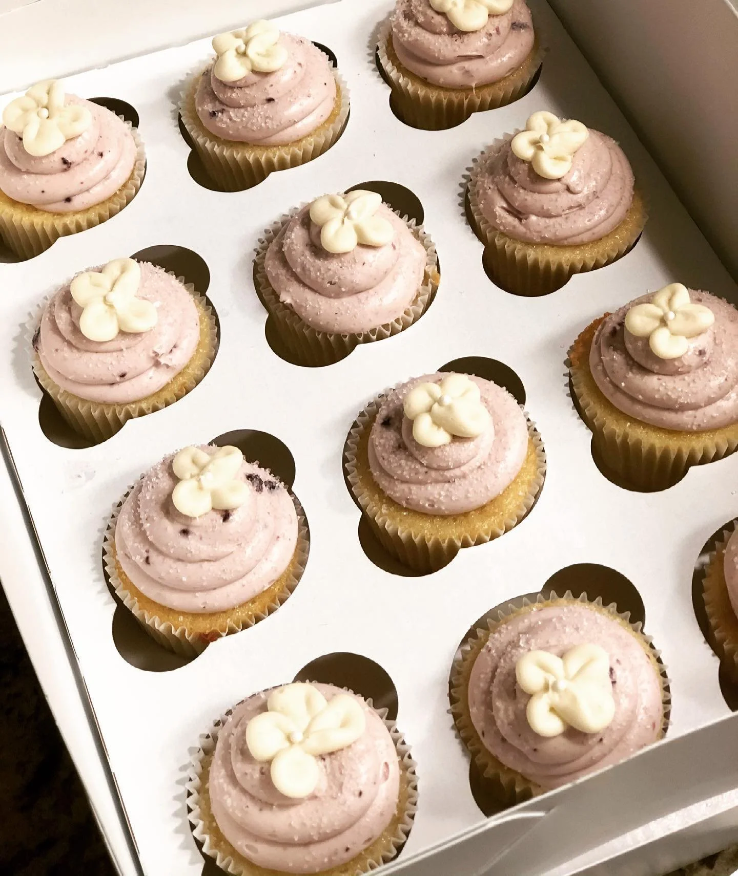 A box of cupcakes with pink frosting, decorated with small vanilla flower-like toppers, arranged in a white box with individual compartments.
