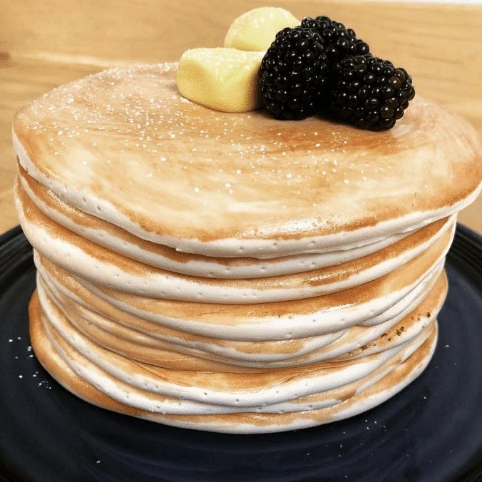 Stack of seven pancakes topped with butter, blackberries, and powdered sugar, served on a dark plate.