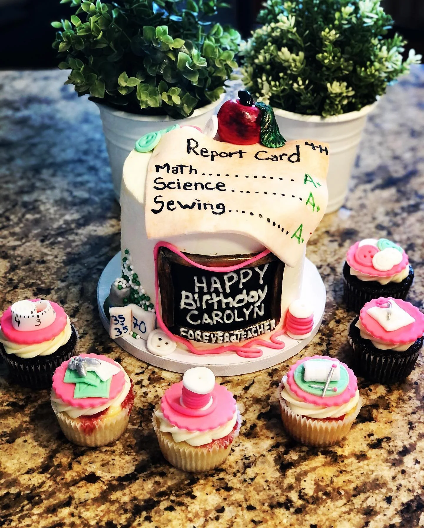 A birthday cake decorated with a report card theme, featuring a list of subjects and grades, surrounded by pink and white cupcakes with school-themed toppers, two potted plants, and a granite countertop.
