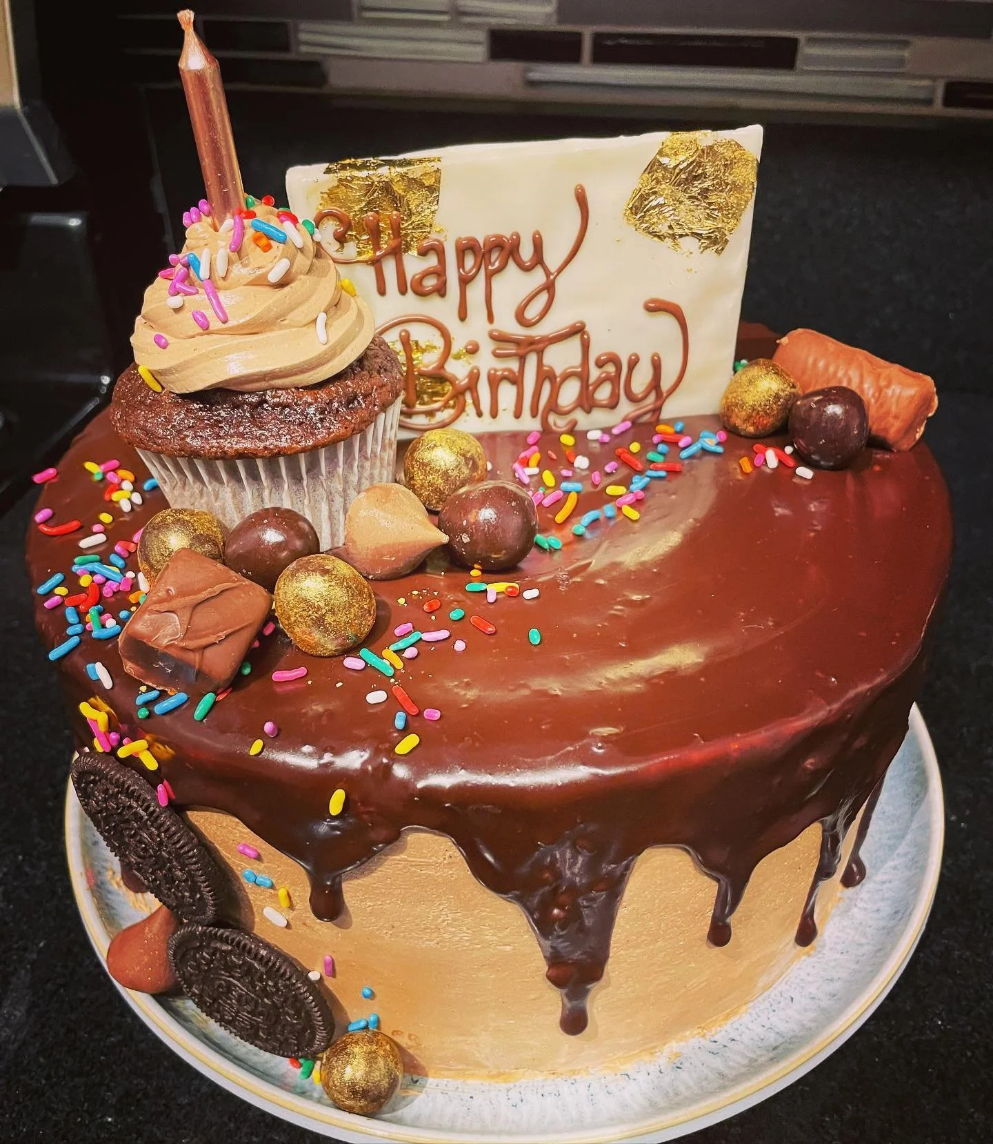 Chocolate birthday cake decorated with colorful sprinkles, gold candies, cookies, chocolates, a cupcake with frosting and a candle, and a white chocolate plaque with red icing that says 'Happy Birthday'.