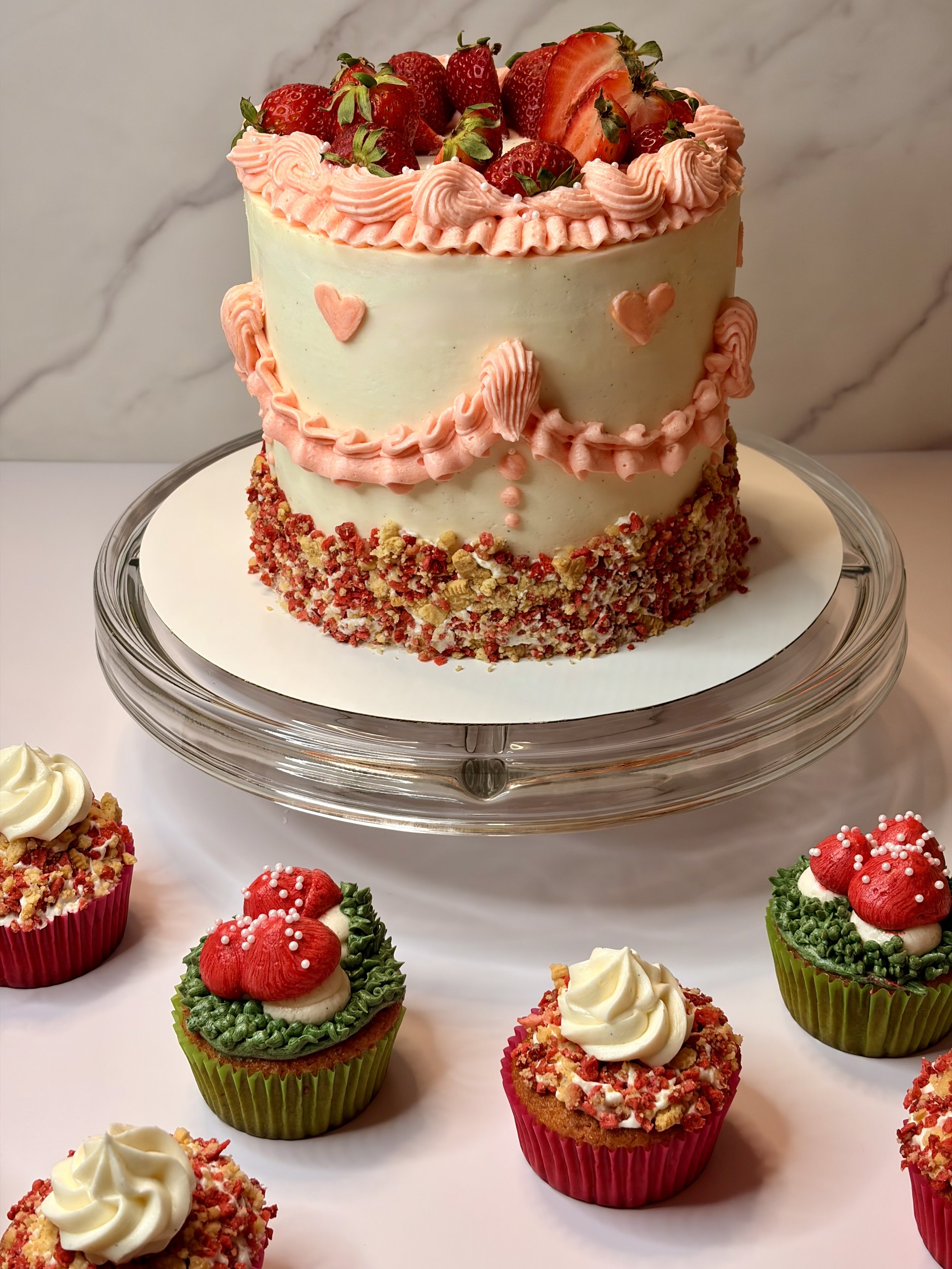 A two-tiered cake decorated with strawberries and pink frosting, and small pink hearts on the side, sitting on a glass cake stand. There are various decorated cupcakes around the cake with strawberries, frosting, and sprinkles.