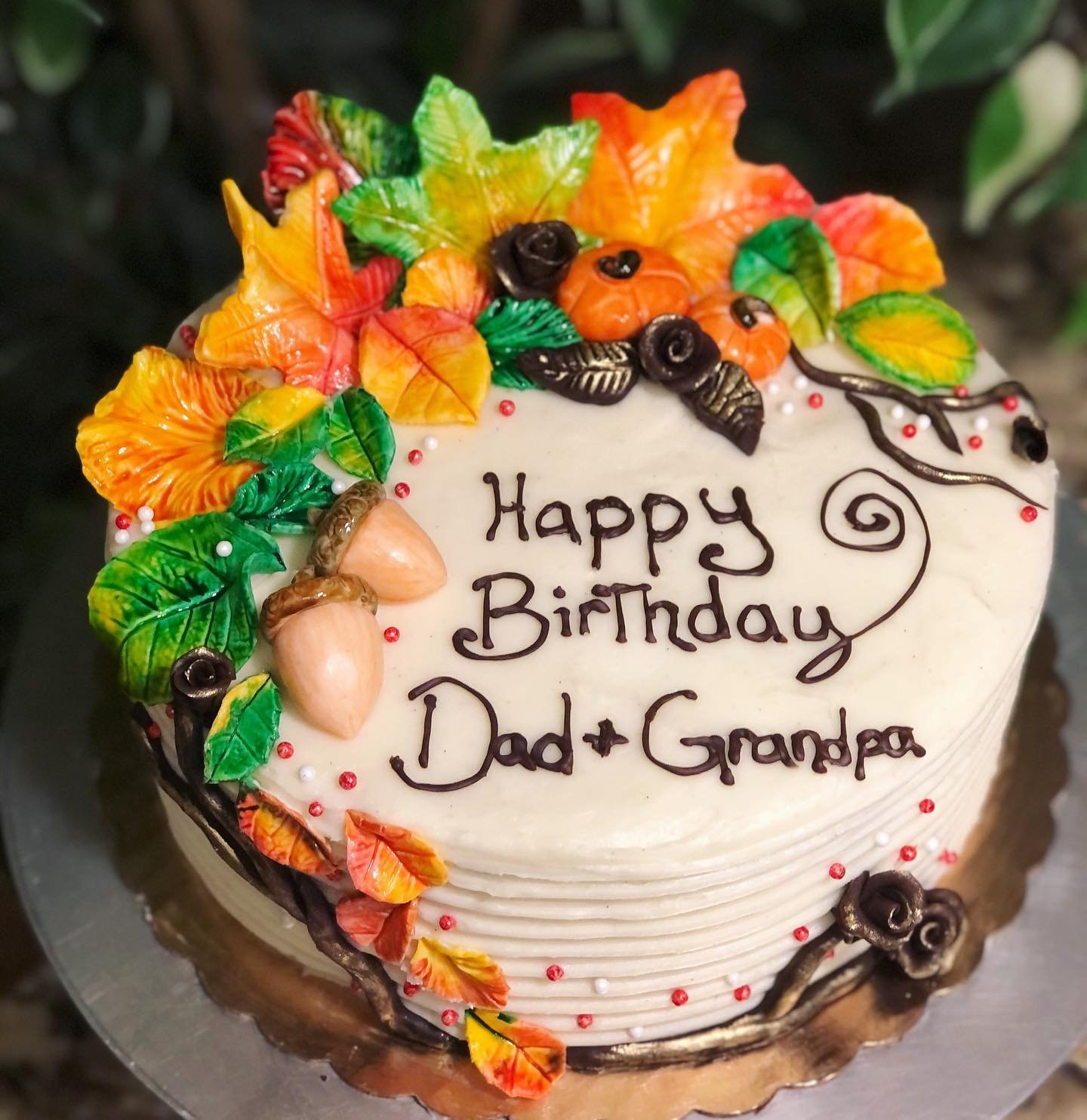 A birthday cake decorated with colorful autumn leaves, small pumpkins, acorns, and berries. The cake has the message 'Happy Birthday Dad + Grandpa' written in chocolate.