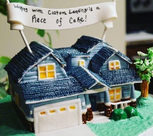 A cake designed to look like a house, with a sign on top that reads "Working with Custom Lending is a piece of cake".