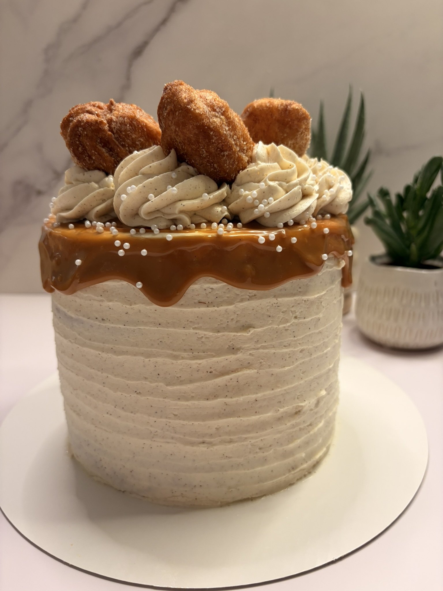 A layered dessert cake with caramel and cream frosting, topped with fried churros and white edible pearls, on a white cake board with a small decorative plant in the background.