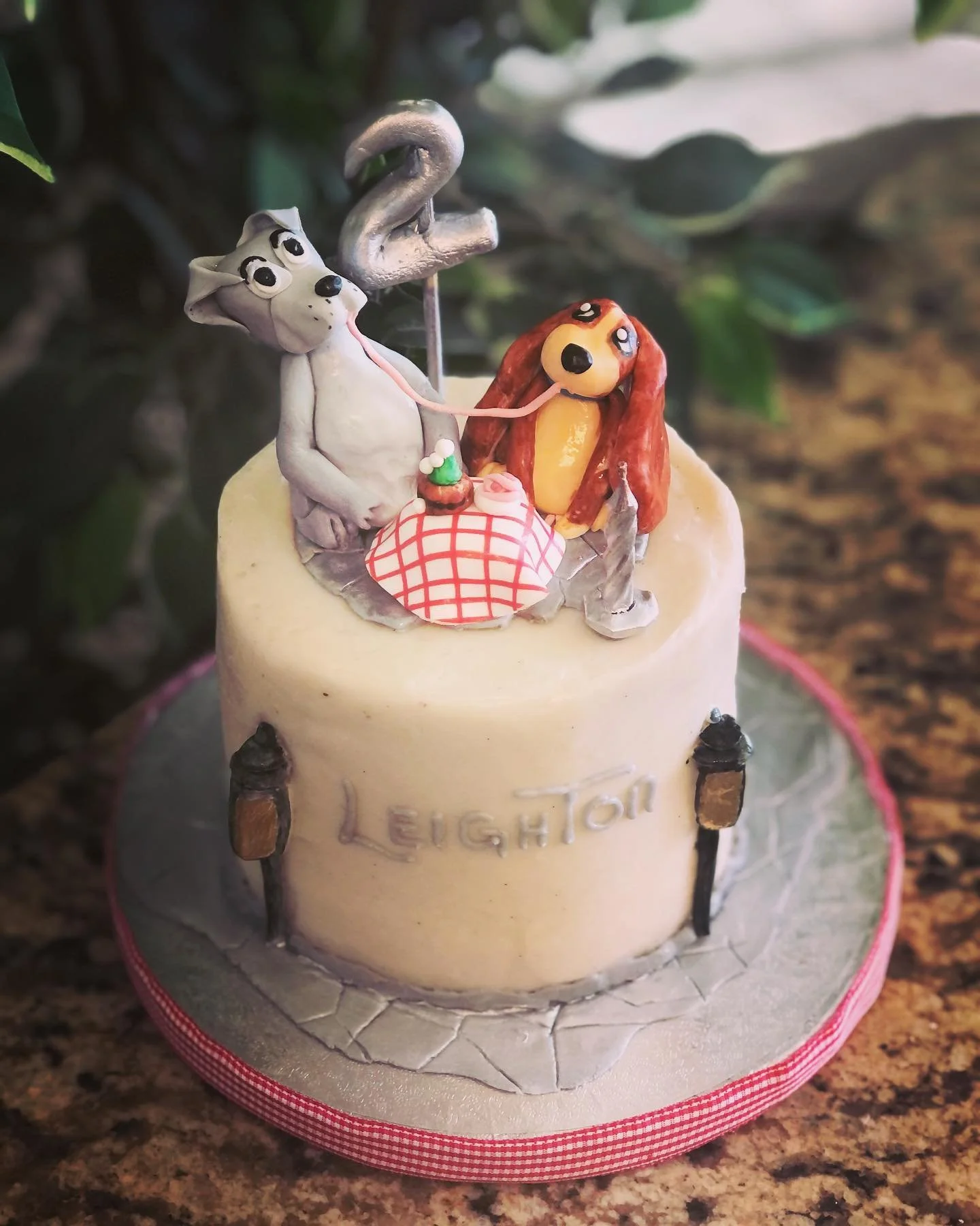Cake topper featuring a white dog and a girl with red hair, sitting at a table with a checkered tablecloth, with the word 'LEIGH' written on the cake.