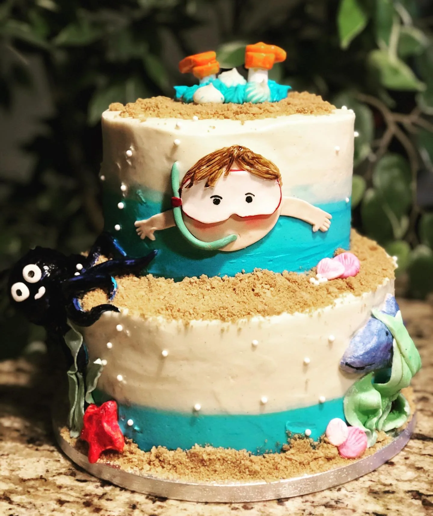A three-tiered birthday cake decorated with an underwater adventure theme, featuring a fondant figure of a kid with a snorkeling mask, an octopus, seashells, and ocean waves.