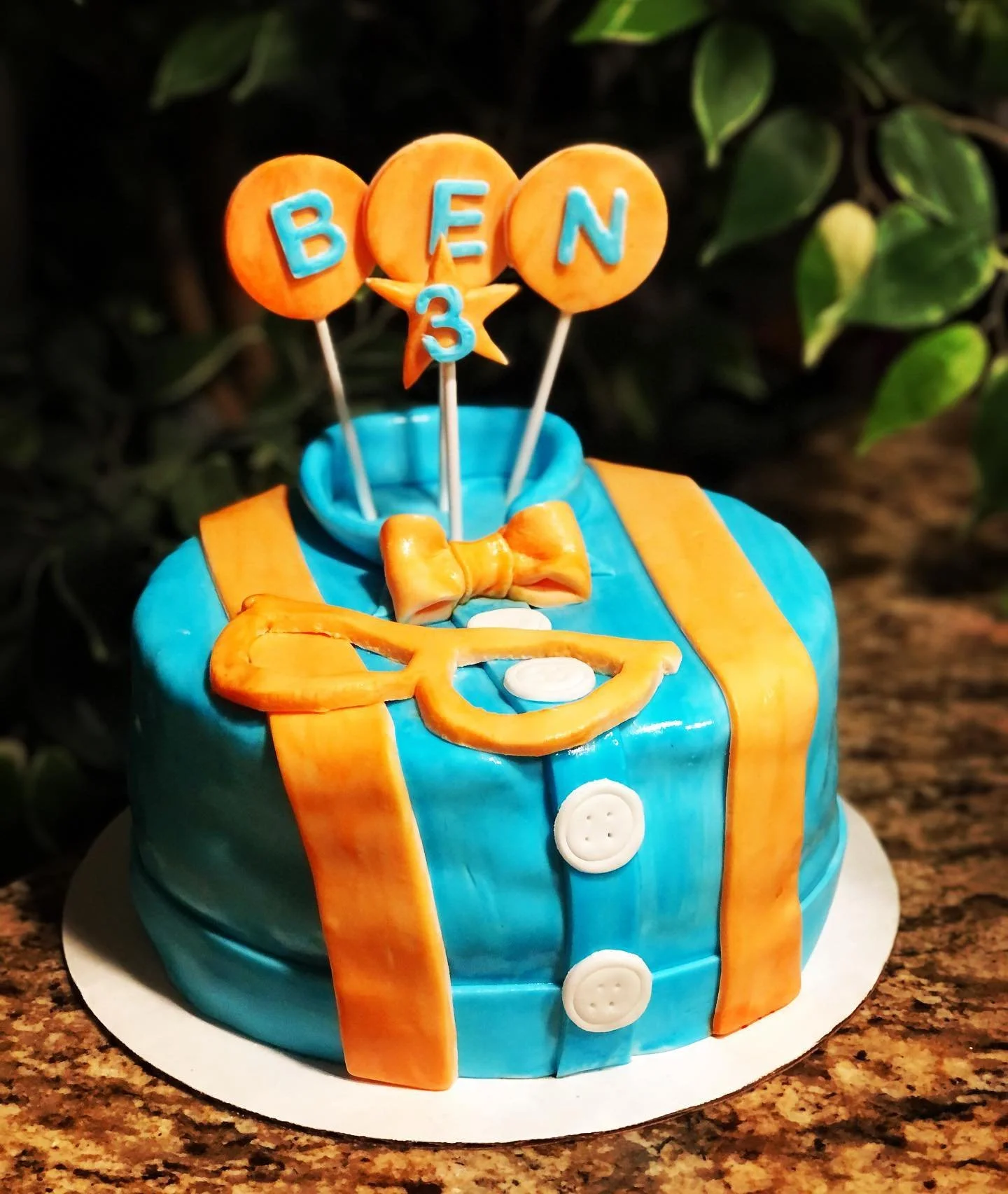 A birthday cake decorated with a blue suit with orange suspenders, a yellow bow tie, and white buttons, topped with orange cookies spelling 'BEN 3' and a star, set against a dark background with green leaves.