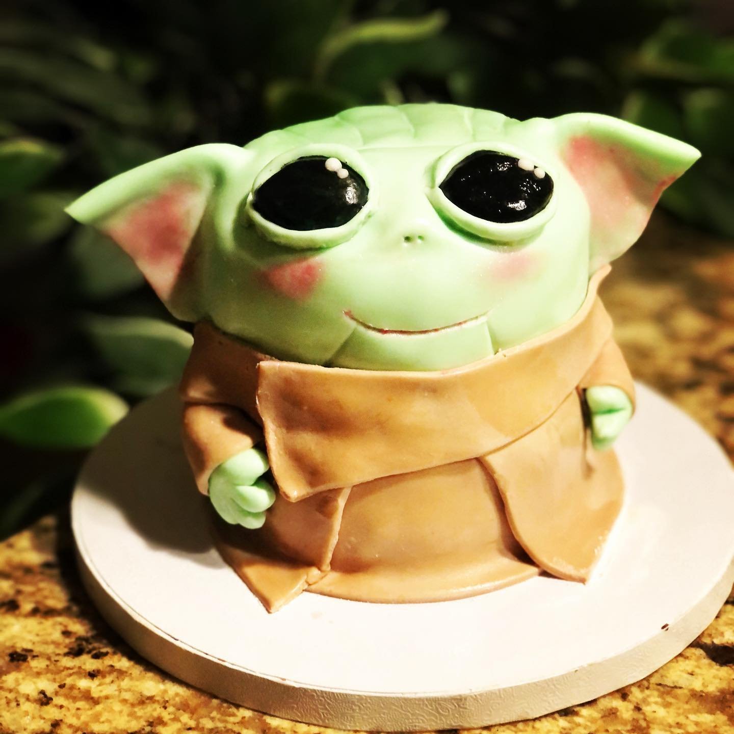 A cake designed to look like Baby Yoda from Star Wars, with large black eyes, green skin, and ears, wearing a brown robe.