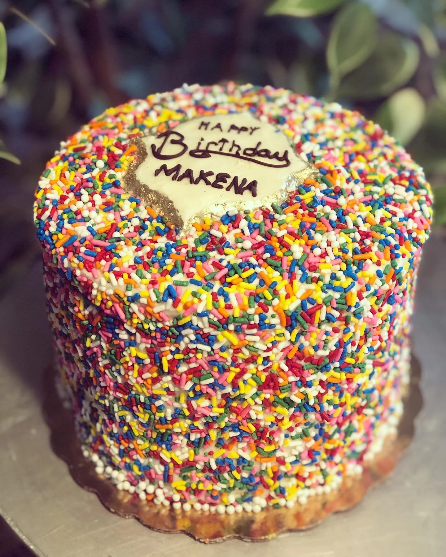 A round birthday cake decorated with colorful rainbow sprinkles covering the entire surface. A white icing plaque with gold edges in the center of the cake has the message 'Happy Birthday Makena' written in purple icing.