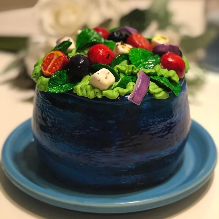 A cake decorated with realistic-looking edible vegetables on top and wrapped in a shiny blue icing, sitting on a blue plate.