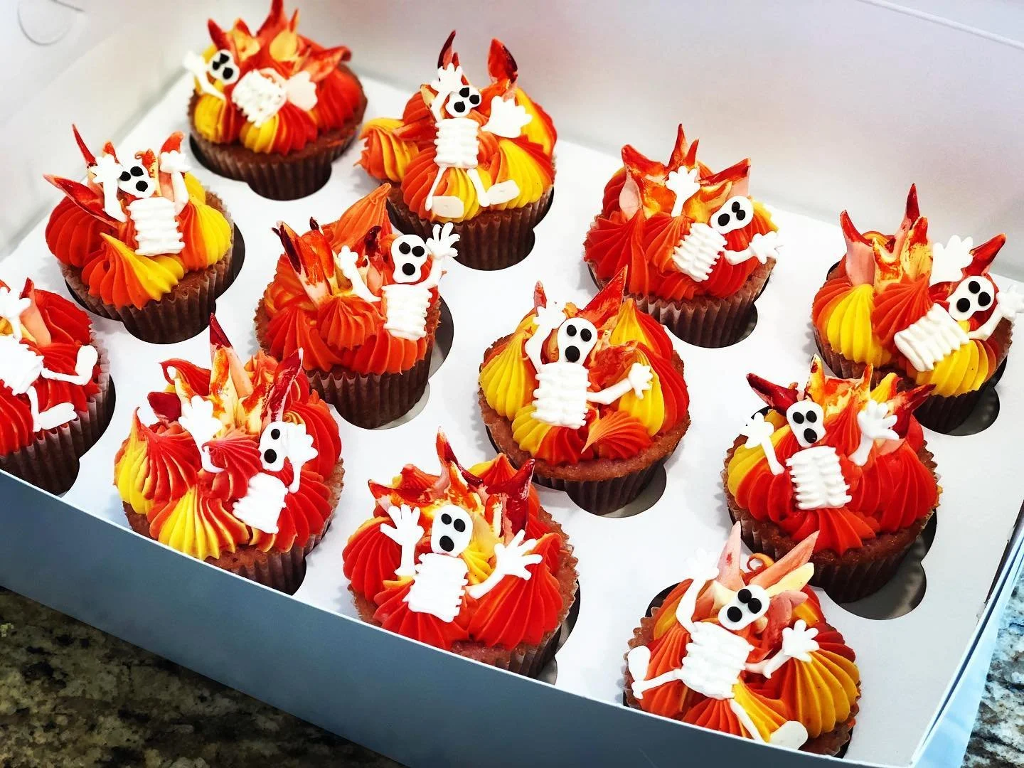 Cupcakes decorated with orange, yellow, and red frosting, with skeleton and ghost themed edible toppers, arranged in a white box.
