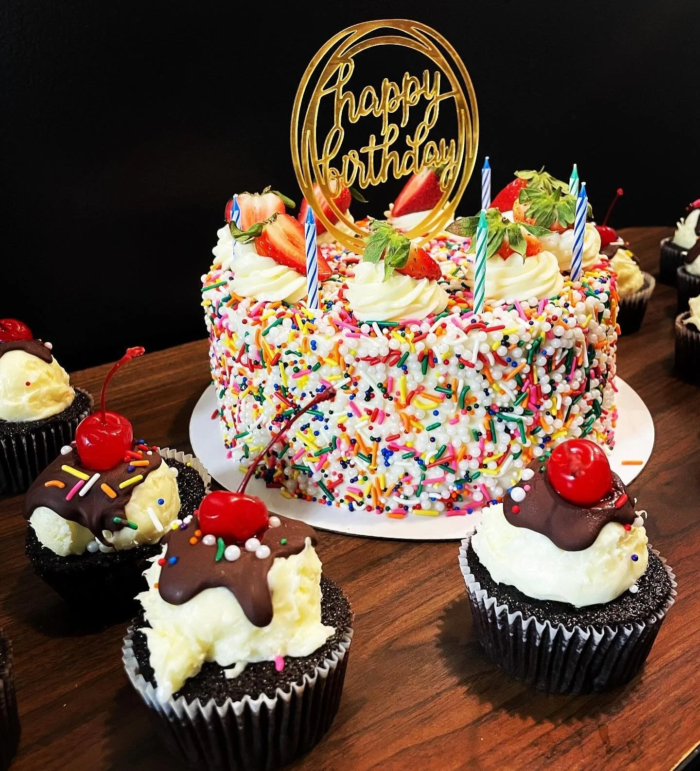 A colorful birthday cake with rainbow sprinkles, strawberries, whipped cream, and a 'happy birthday' topper. Surrounding cupcakes with cherries, chocolate, and sprinkles are on a wooden table.