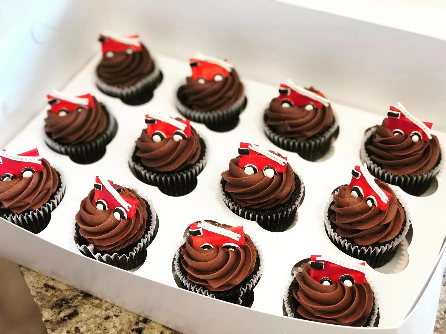 Box of 12 chocolate cupcakes decorated with fire truck toppers.