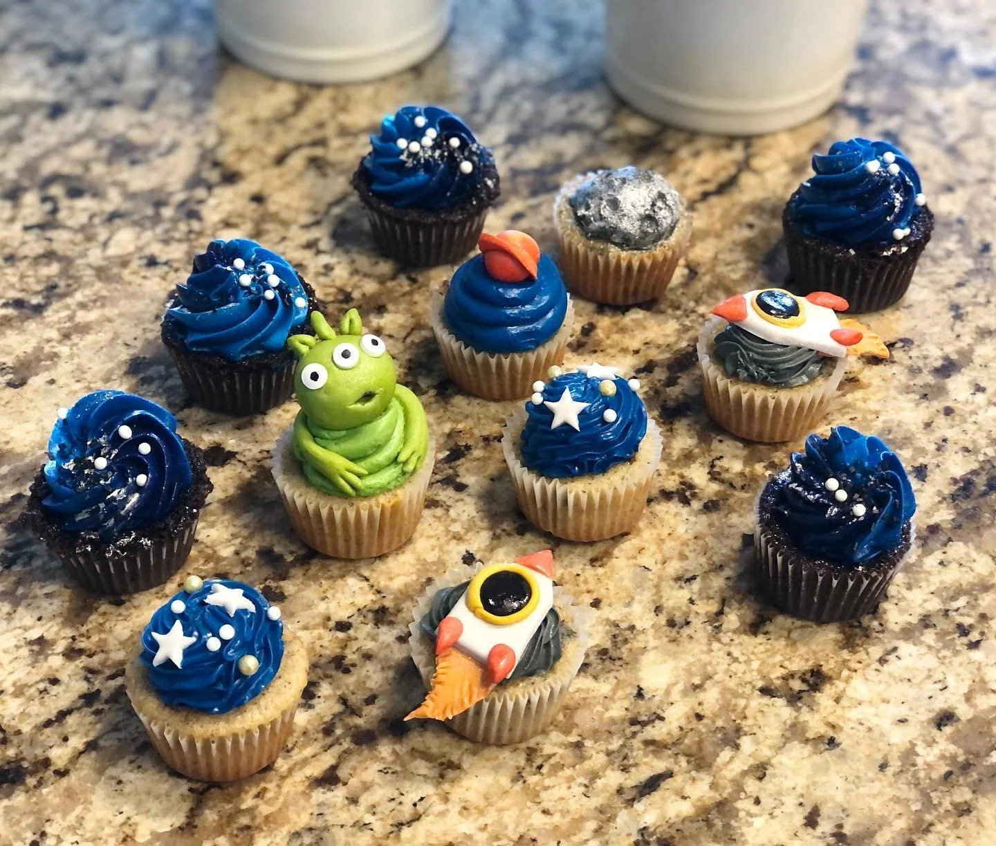 Assorted space-themed decorated cupcakes on a granite countertop. Some cupcakes feature blue frosting with white sprinkles, one has a green alien figure, and others are decorated with rockets, stars, and planets.