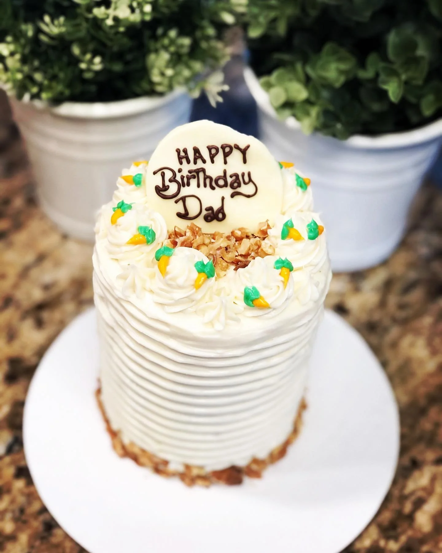 A birthday cake with white frosting, colorful sprinkles, and a white chocolate disc with "Happy Birthday Dad" written on it, topped with chopped nuts.