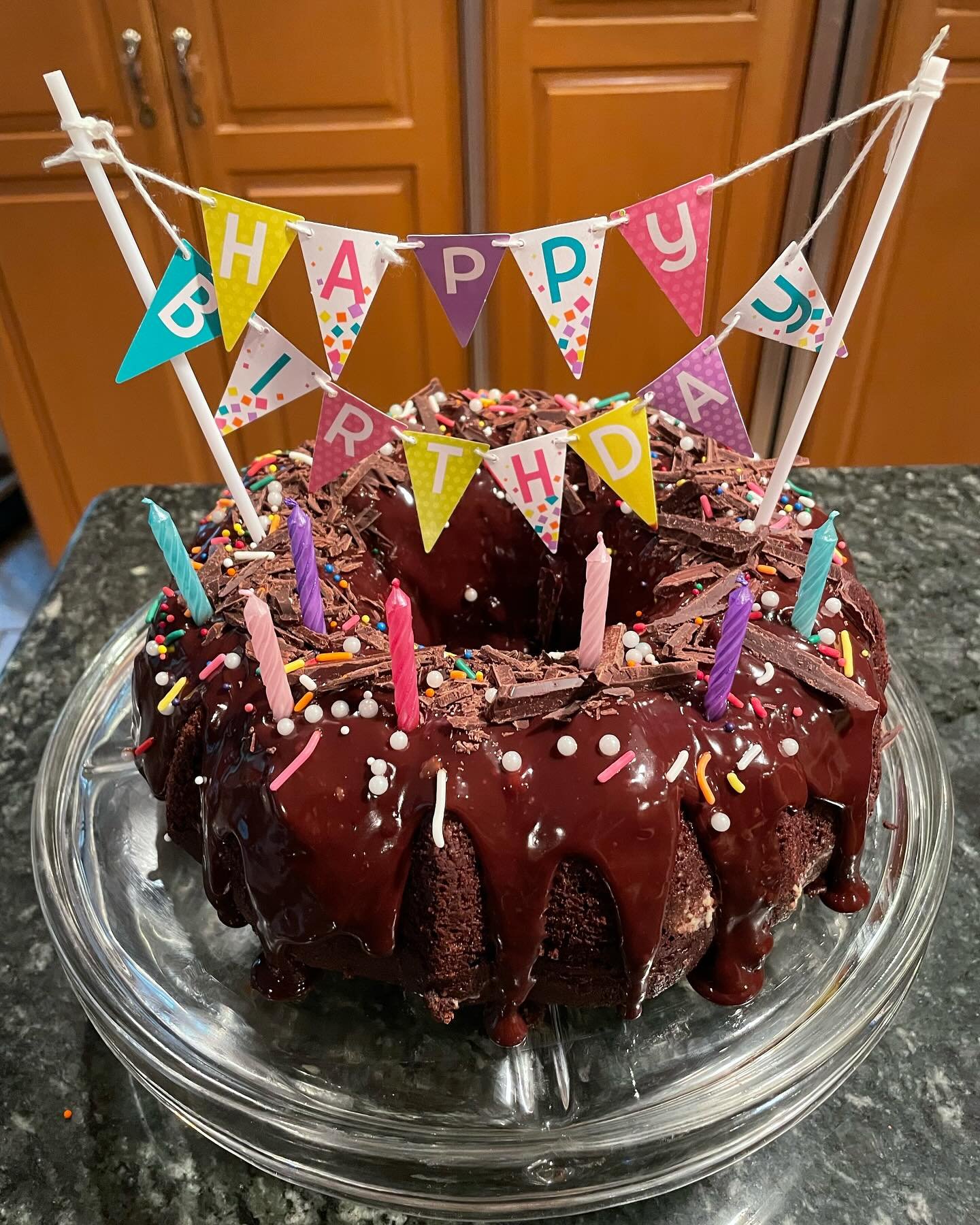 Chocolate cake with chocolate shavings, colorful sprinkles, and chocolate ganache, topped with pink, purple, and blue candles, with a banner that reads 'Happy Birthday' in colorful letters.