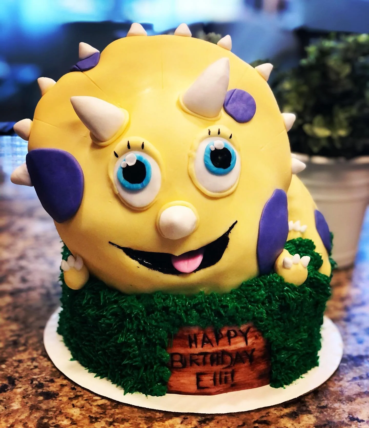 A cake designed as a cartoon creature with a round yellow body, big blue and black eyes, white spikes on its head, purple spots on its body, and a smiling face, sitting on a green grassy base with a birthday message.