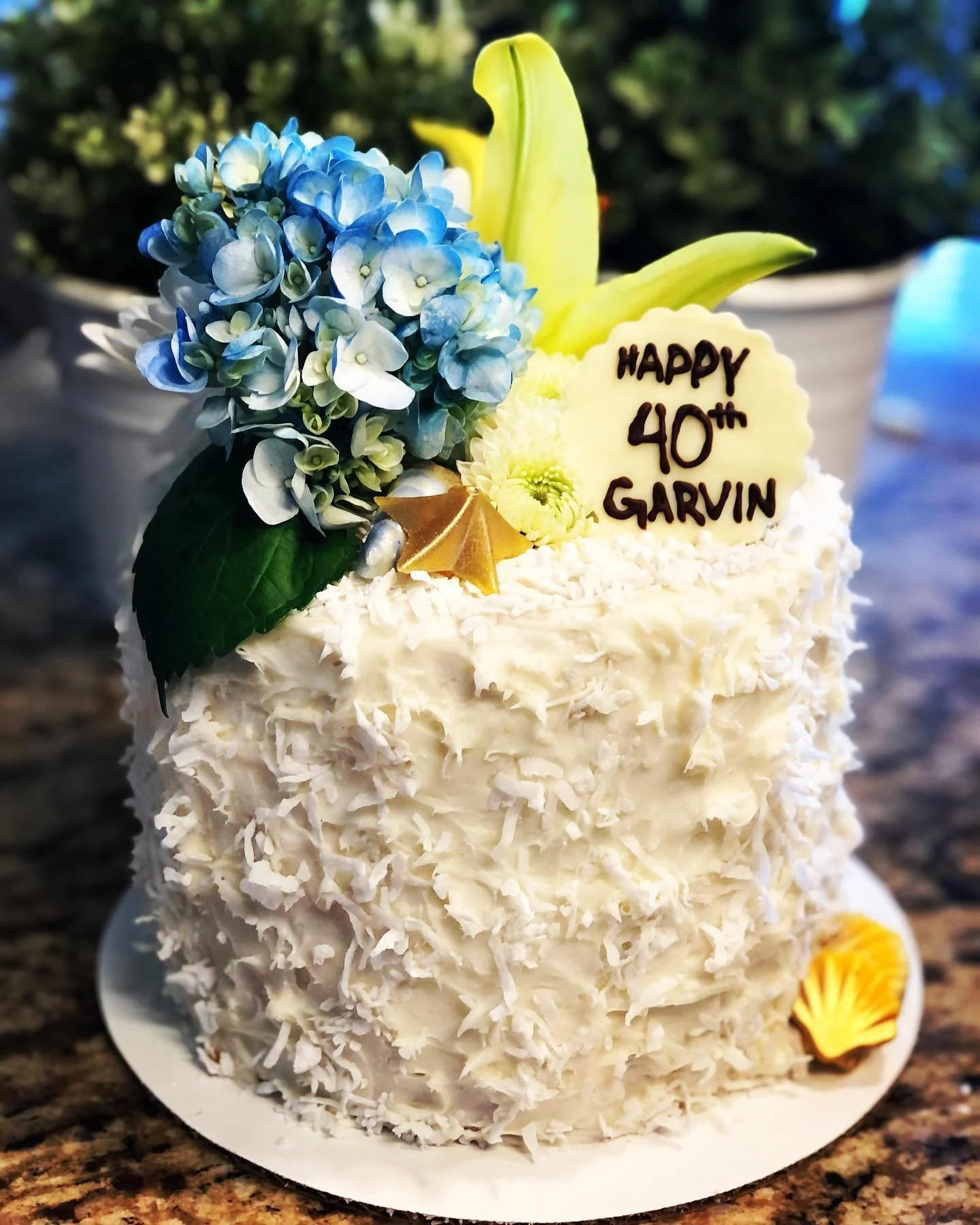 A tall birthday cake covered with white shavings, decorated with a bouquet of blue and white flowers, a yellow lily, a white chocolate plaque with "Happy 40th Garvin" written in dark chocolate, and small decorative items.