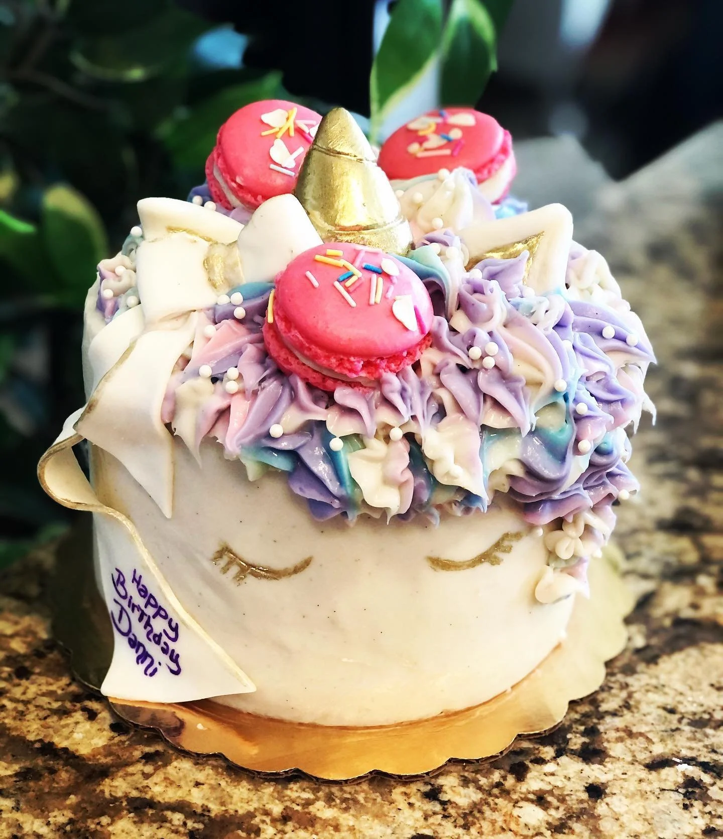 A birthday cake decorated with pastel purple and blue frosting, topped with pink macarons, a gold unicorn horn, white whipped cream, and colorful sprinkles.
