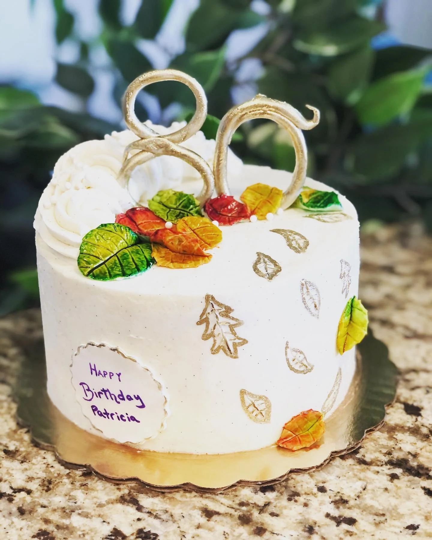 A white birthday cake decorated with colorful edible leaves, gold leaf accents, and two decorative curly white sticks on top. A small note on the side reads, 'Happy Birthday Patricia'.