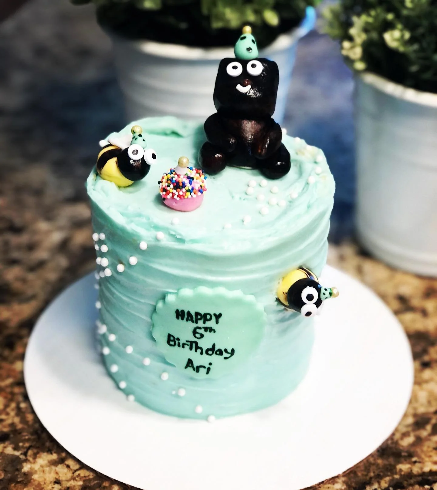 A decorated birthday cake with mint green frosting, featuring small white pearl-like decorations, a message that reads 'HAPPY 6TH Birthday Ari,' and various bee-themed fondant decorations including bees and a hive topped with colorful sprinkles. Ther