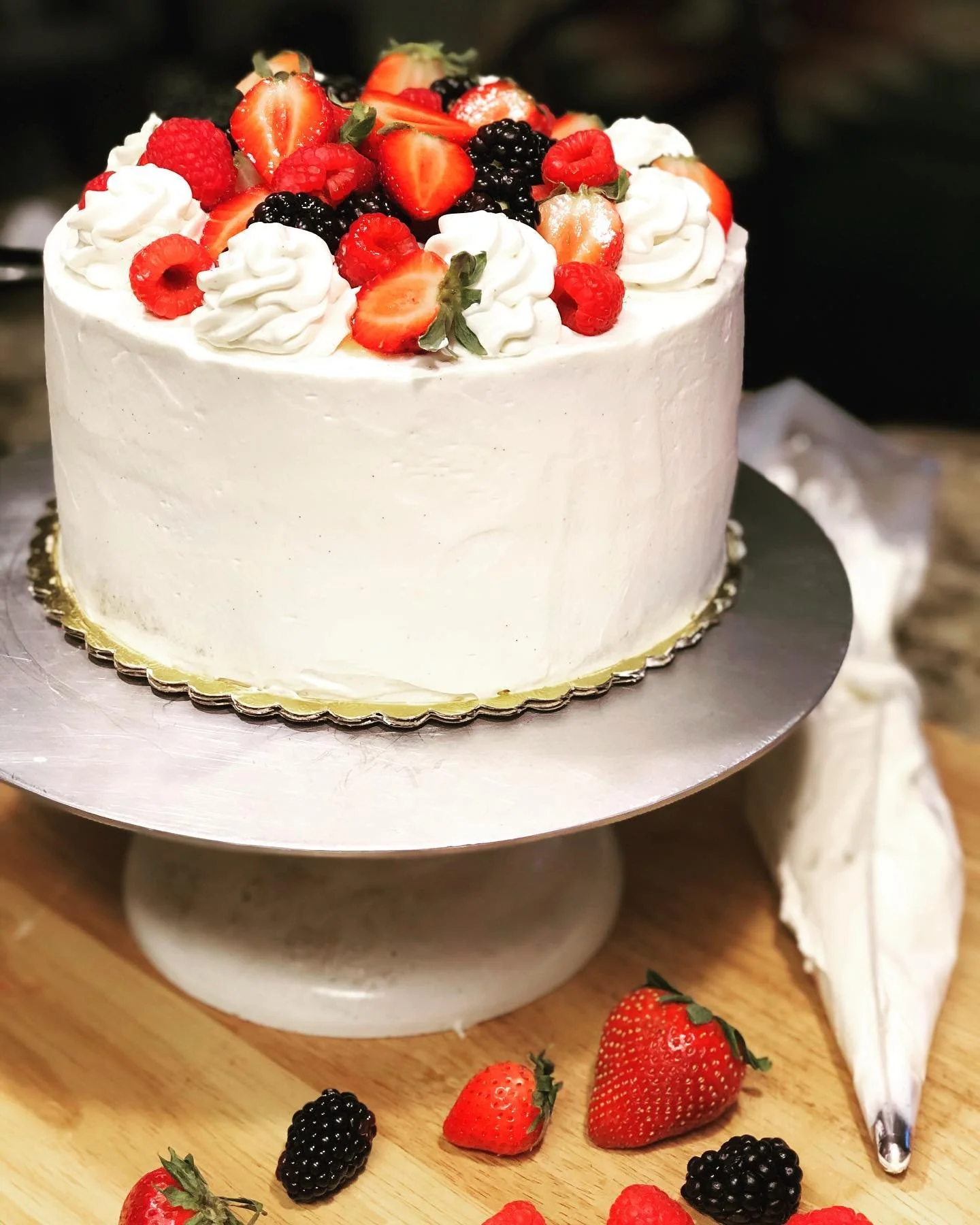 A white frosted cake topped with fresh strawberries, blackberries, and raspberries, on a silver cake stand with scattered berries and strawberries on a wooden surface.