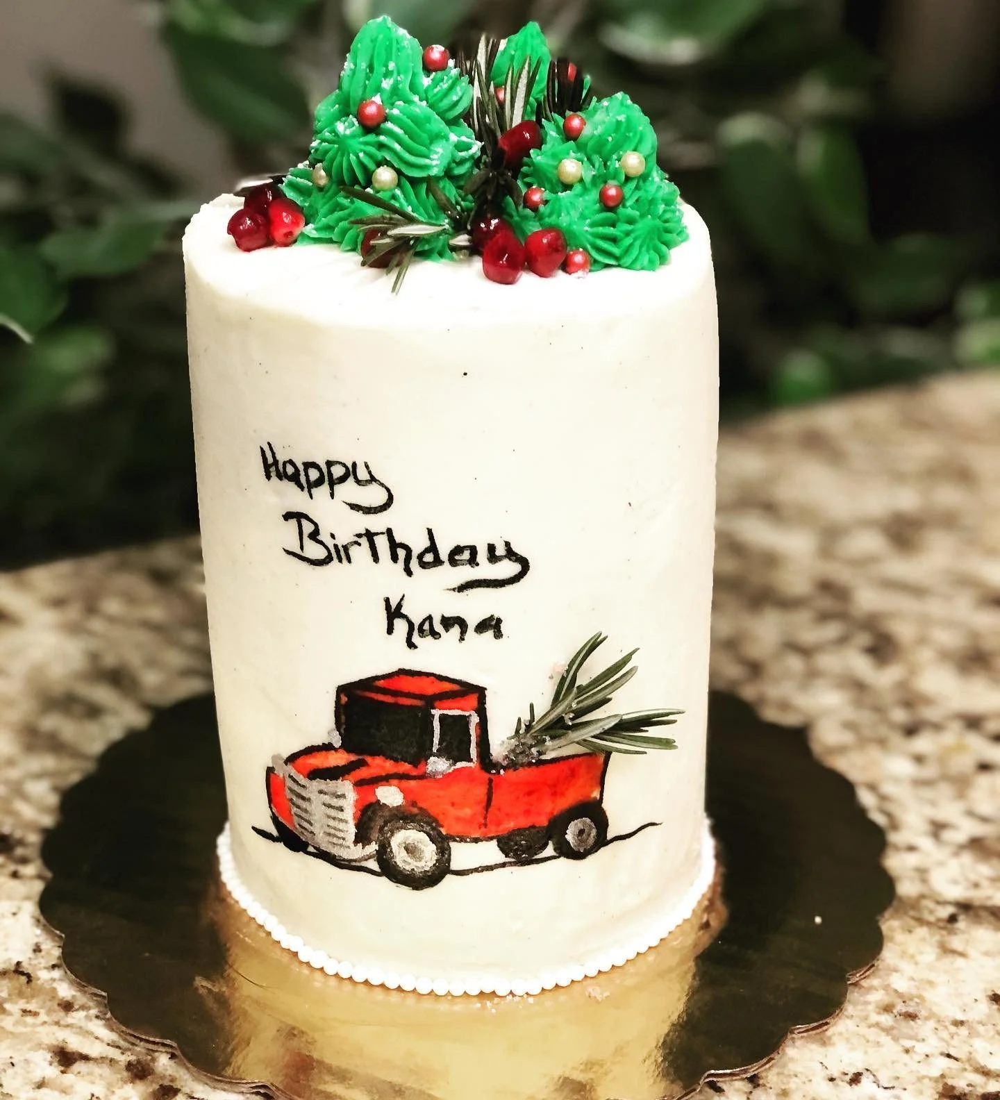A white birthday cake decorated with mini green icing Christmas trees, red berries, and small pearl-like ornaments, featuring a hand-drawn red truck with a Christmas tree in the back and the message 'Happy Birthday Kana' written on the front.