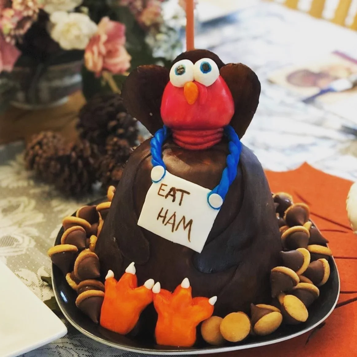 Decorative turkey-shaped cake with a red candy face, big eyes, orange beak, and blue necklace, holding a sign that says "EAT HAM," surrounded by candy candy corn and candy chocolates, with a floral and pinecone background.