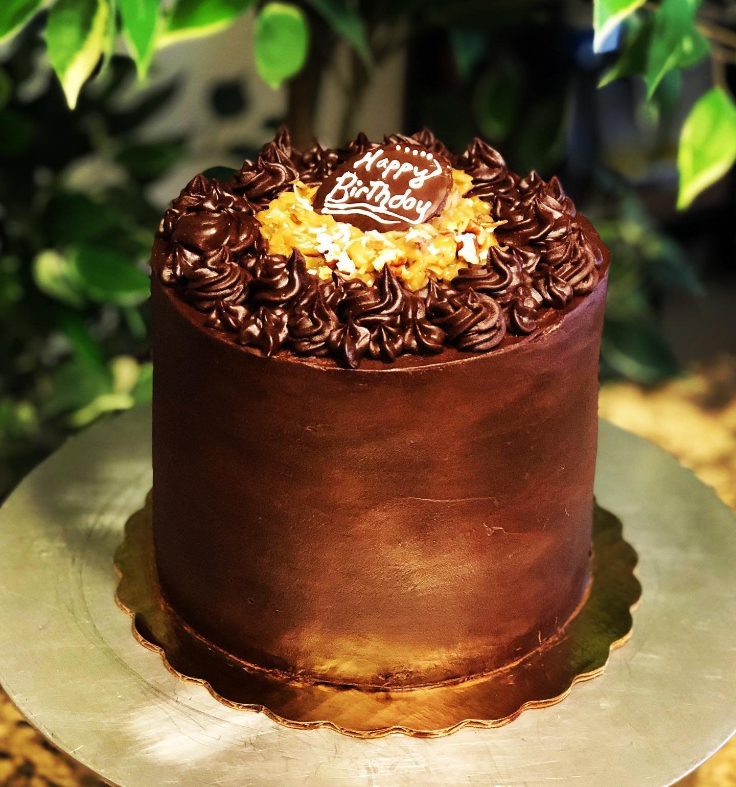 A tall chocolate birthday cake with a smooth chocolate frosting, decorated with chocolate piped rosettes on top, and a chocolate plaque with "Happy Birthday" written on it, surrounded by small white decorations.