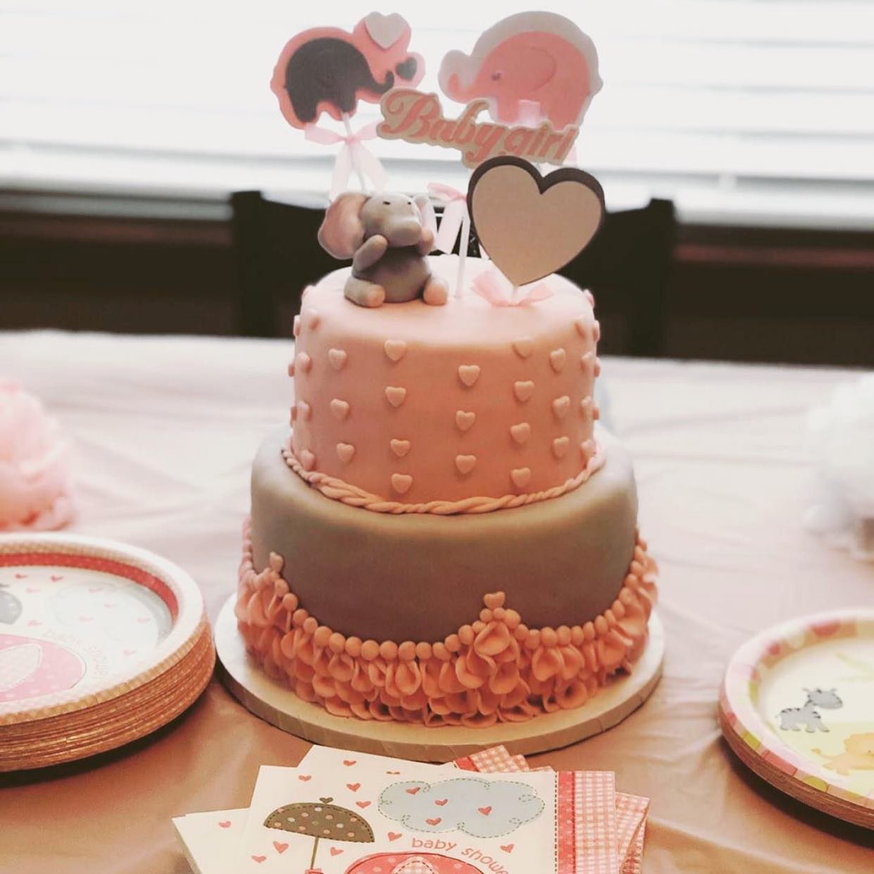 A two-tiered pink and gray cake decorated with small hearts and a rope border, topped with a 'Twinkle' sign, a pink heart, a baby elephant figure, and a gray bunny figure. Plates with baby-themed designs and printed baby shower cards are around the c