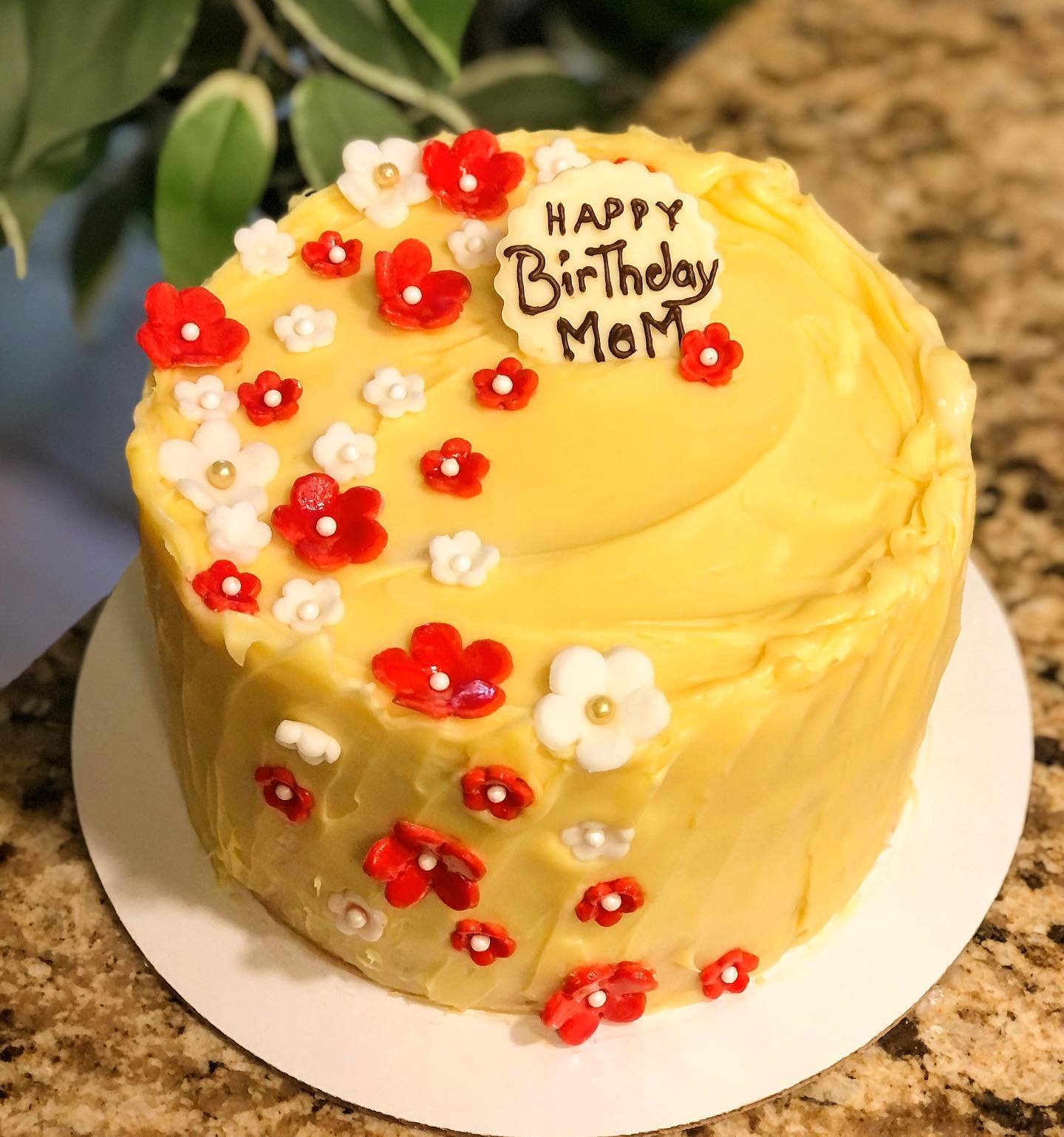 Yellow birthday cake decorated with red and white edible flowers and a message reading "Happy Birthday Mom" written with chocolate, placed on a white cake board on a granite countertop.