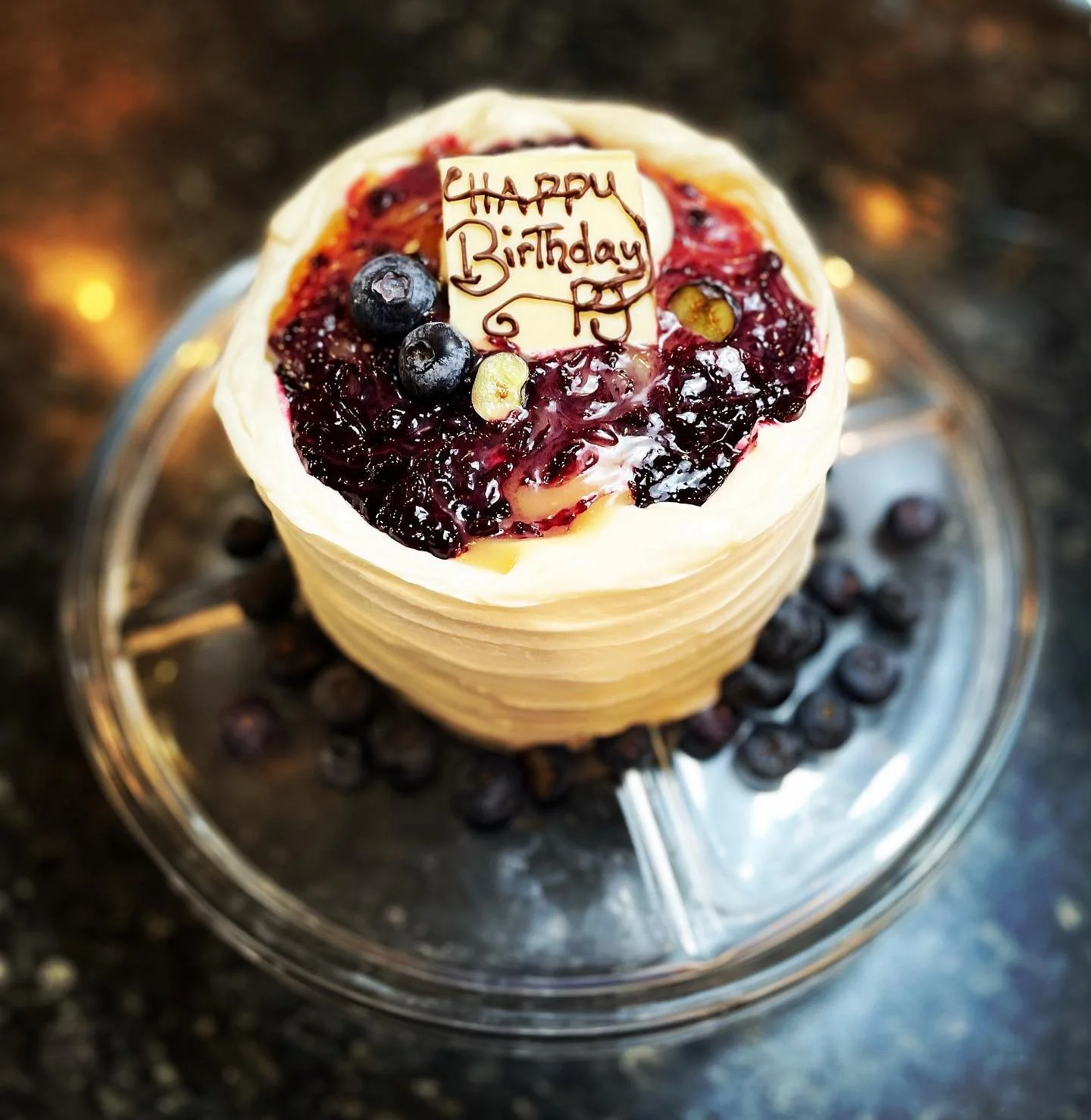 A birthday cake topped with a message that says 'Happy Birthday R' written in chocolate, with blueberries and a berry glaze, placed on a reflective silver tray.