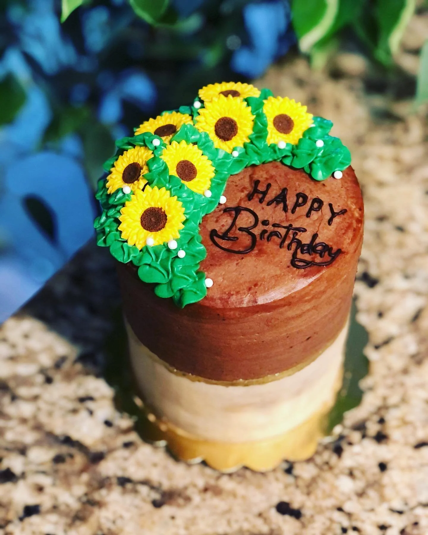 Decorative cake with sunflower frosting and a 'Happy Birthday' message written in black on the top layer, set on a light-colored wooden surface.