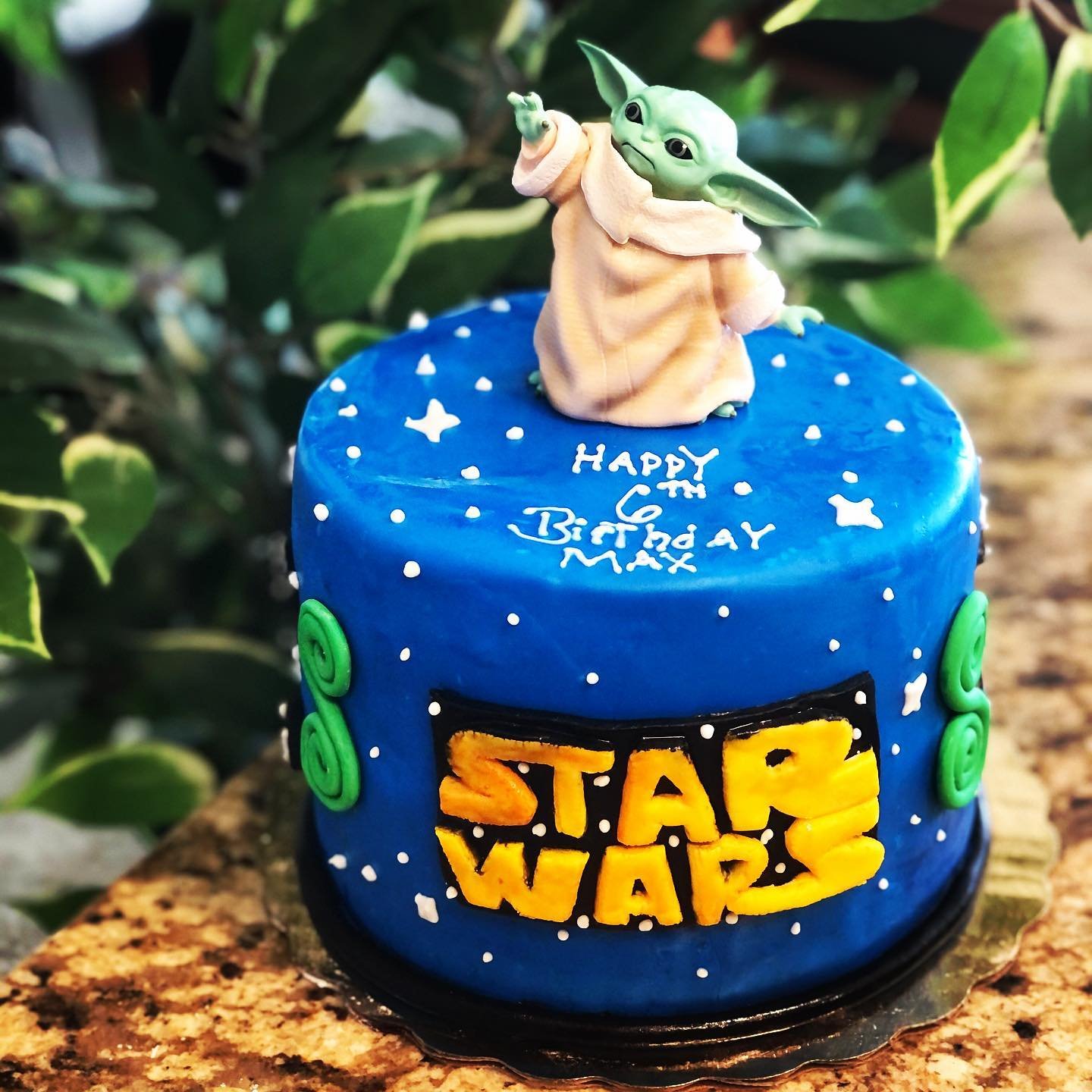 Star Wars themed birthday cake with a Baby Yoda (Grogu) figurine on top, decorated with stars and planets, with the message 'Happy 6th Birthday Max' written on it.