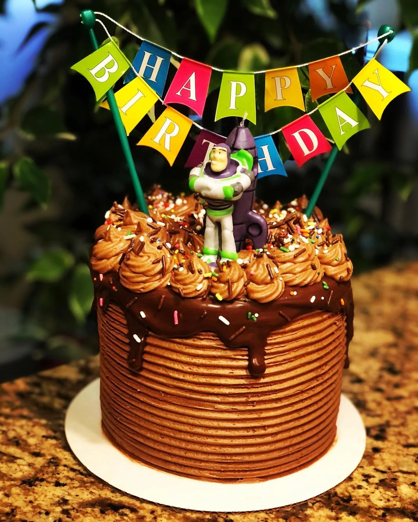 Chocolate birthday cake with whipped frosting, colorful sprinkles, a "Happy Birthday" banner, and a Buzz Lightyear toy figurine on top.