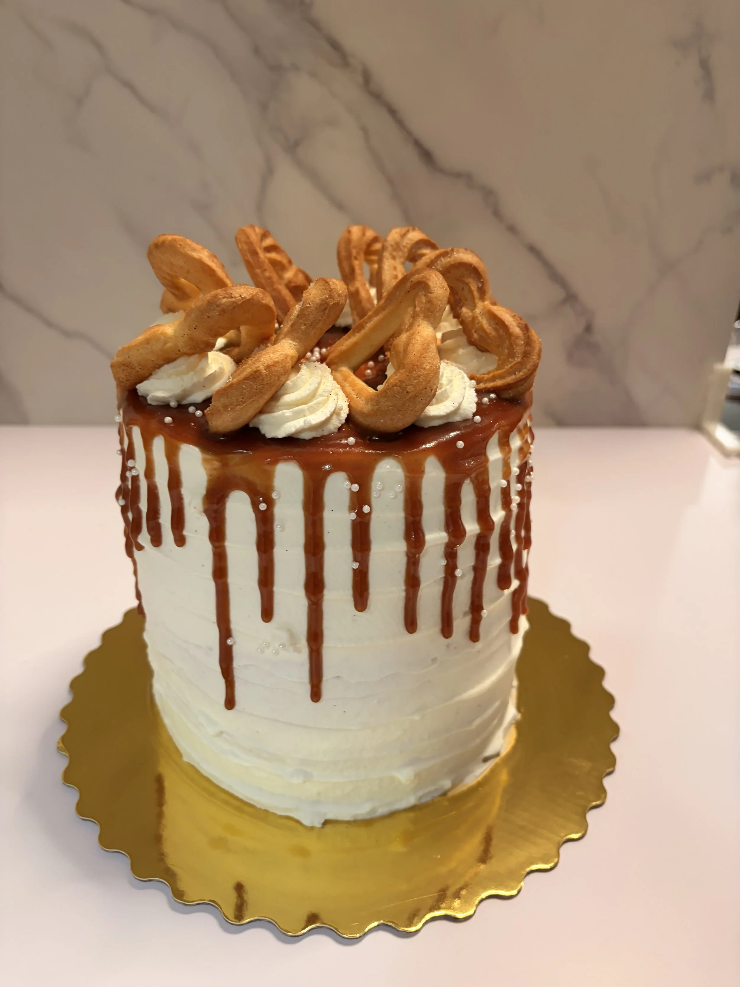 A tall, round birthday cake with white frosting, caramel drip decoration around the edges, topped with cream swirls, and churros arranged on top, placed on a gold cake board.