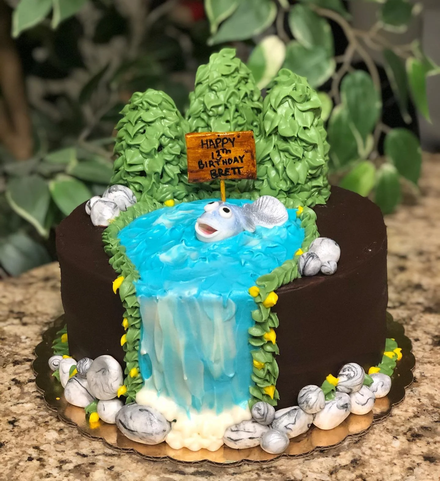 A birthday cake decorated with a woodland scene featuring a waterfall, green trees, and a fish. There is a small wooden sign that reads: 'HAPPY 13th BIRTHDAY BRETT.'