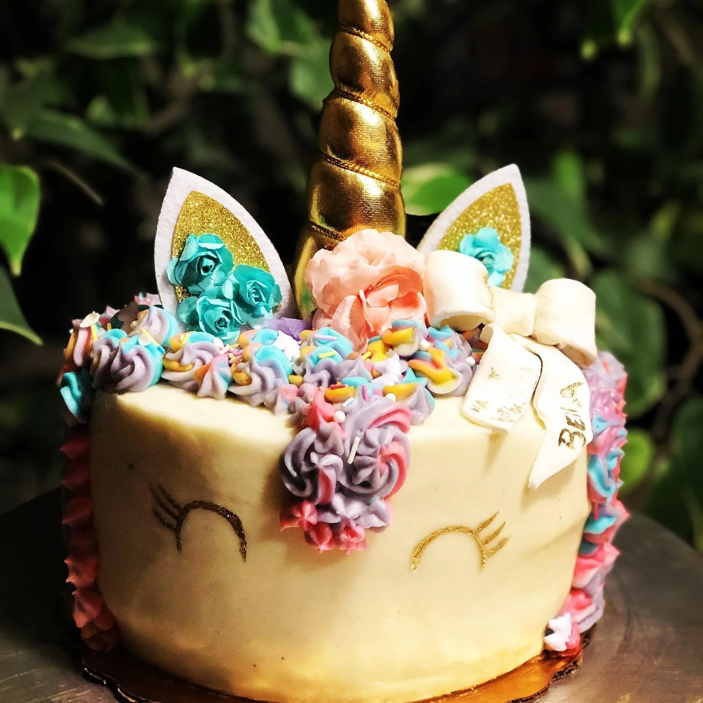 A unicorn-themed birthday cake decorated with a golden unicorn horn, colorful buttercream flowers, sprinkles, and unicorn ears with gold accents.