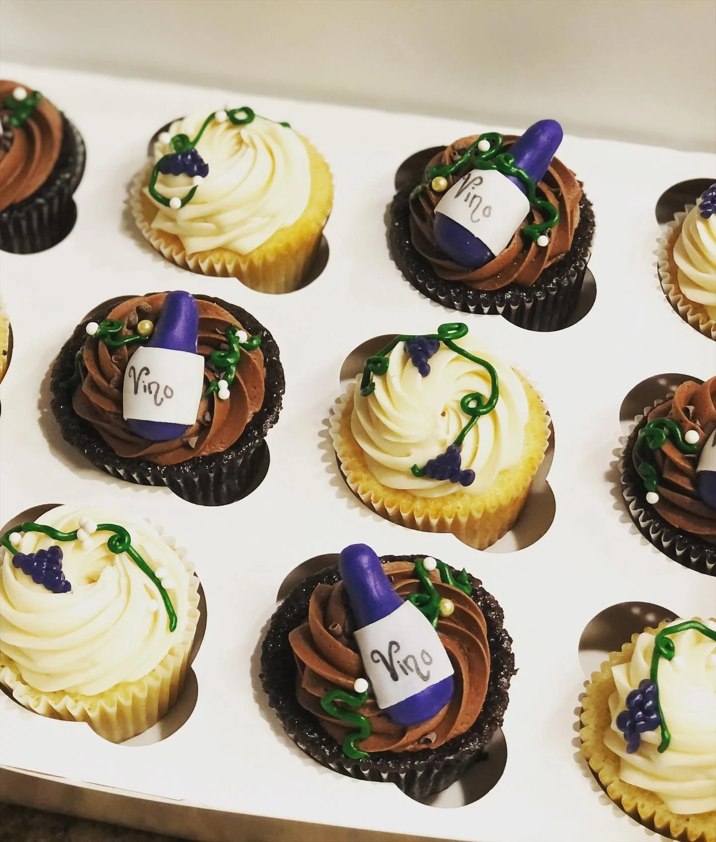 Assorted cupcakes decorated with white and yellow frosting, purple and green edible decorations, and cupcake liners in black and yellow. Some cupcakes feature a small white sign with the word 'Vino' and a purple wine bottle decoration.