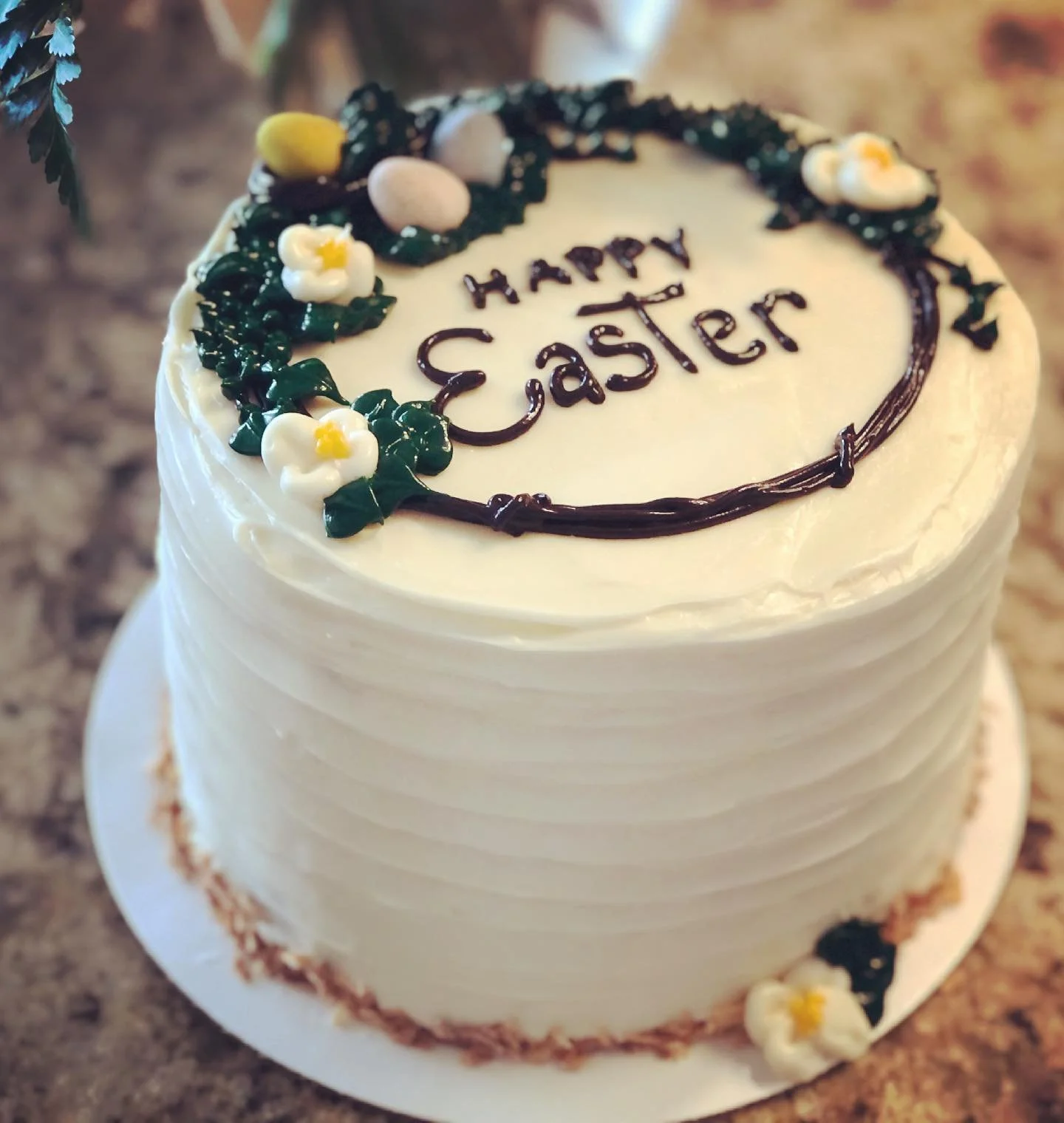 A round white Easter cake decorated with icing flowers and holly leaves, with the words 'Happy Easter' written in the center.
