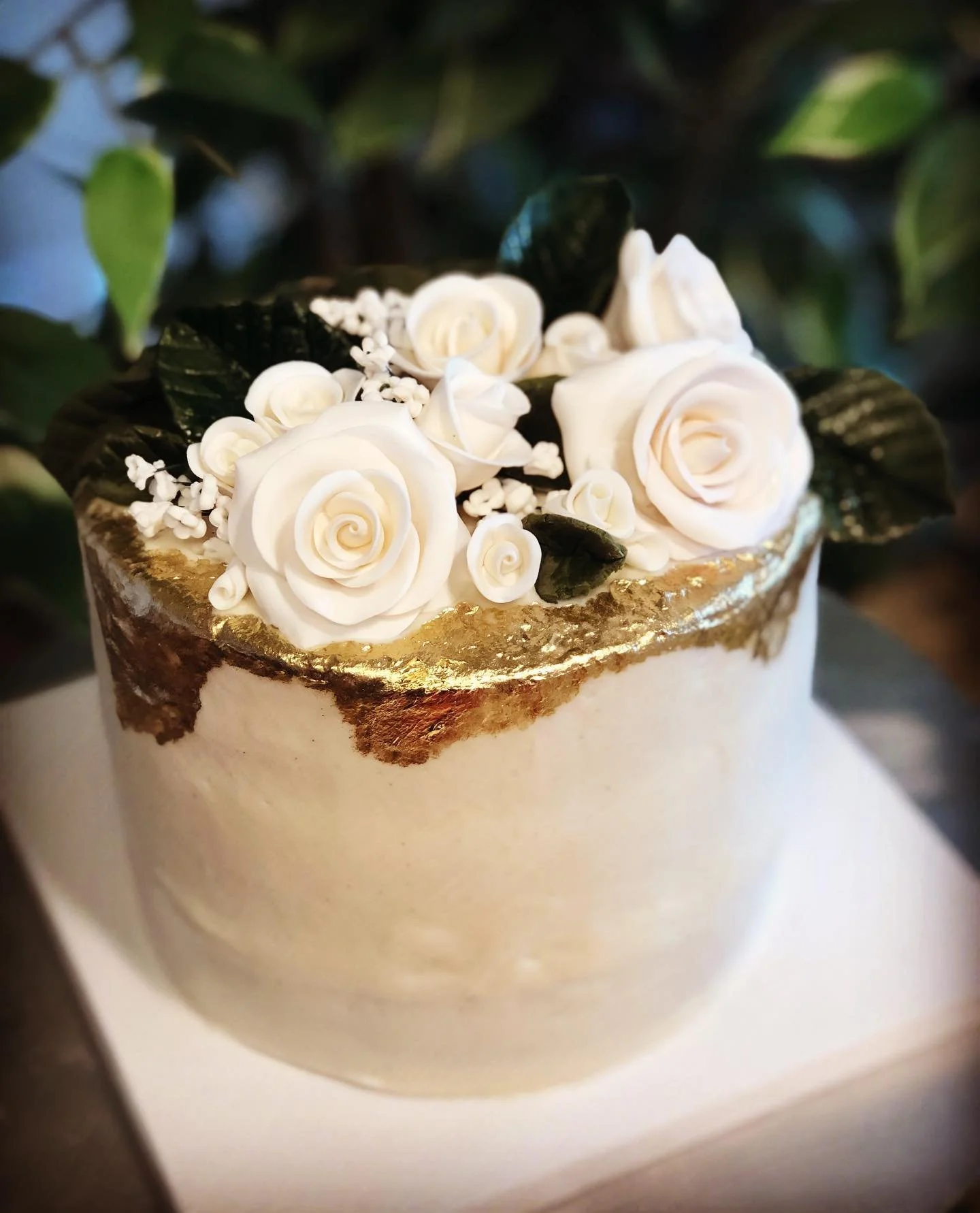 A decorative cake with white roses and green leaves on top, gold foil accents around the edge, and a white base.