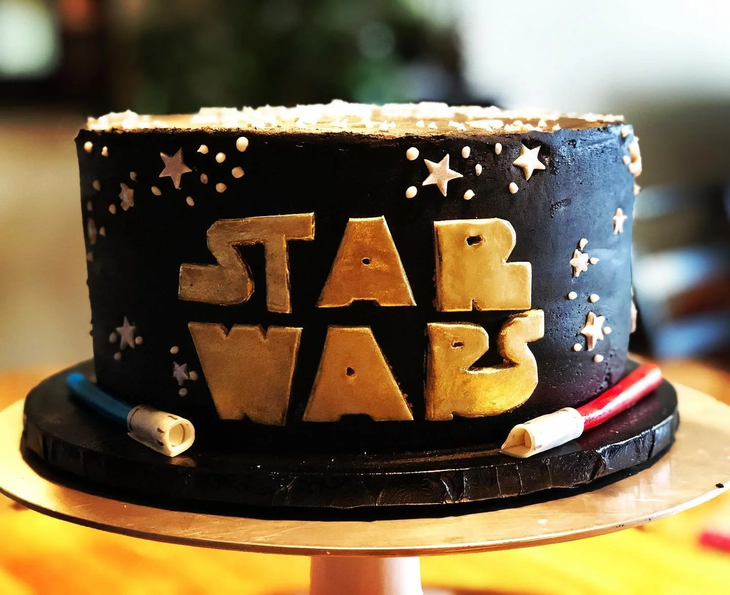 Star Wars-themed cake with black frosting, gold 'Star Wars' lettering, white star and dot decorations, and two lightsaber toppers at the base.