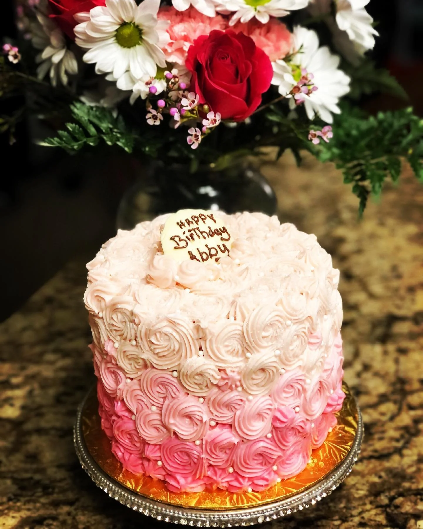 A pink and white rosette cake with a heart-shaped cookie on top that has the message "Happy Birthday Abby" written in chocolate. In the background, there is a bouquet of white, pink, and red flowers with green leaves.