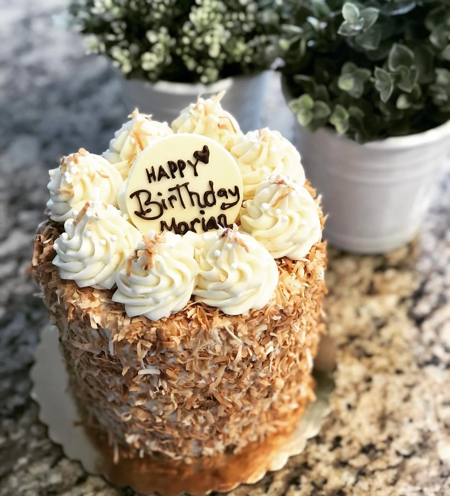 A birthday cake with white frosting and shredded coconut coating, topped with piped cream and a white chocolate disc that says 'Happy Birthday Marion' in chocolate, with a small heart.