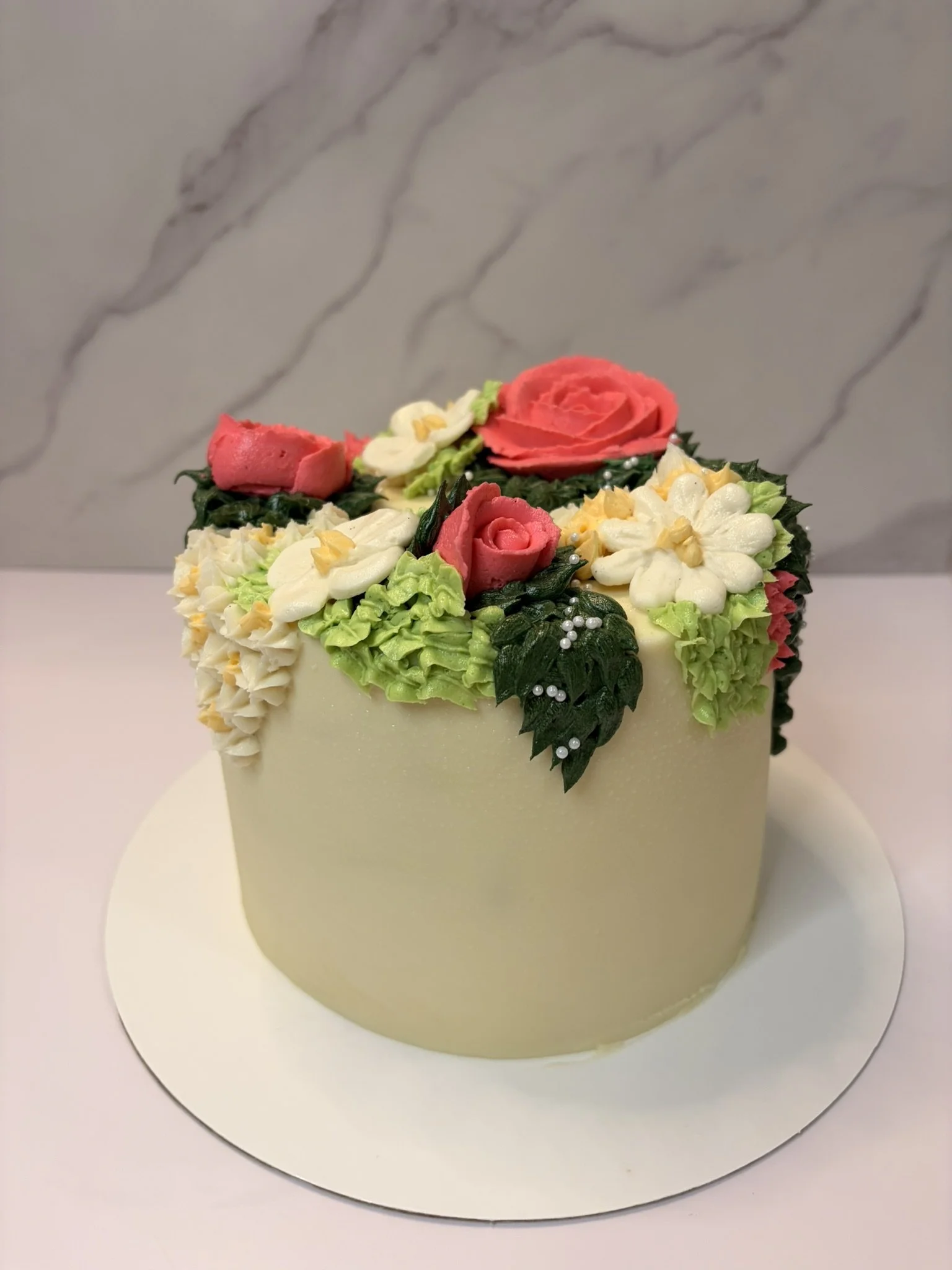 A white cake decorated with colorful buttercream flowers and roses, with green and dark leaves, placed on a white cake board against a marble-patterned background.
